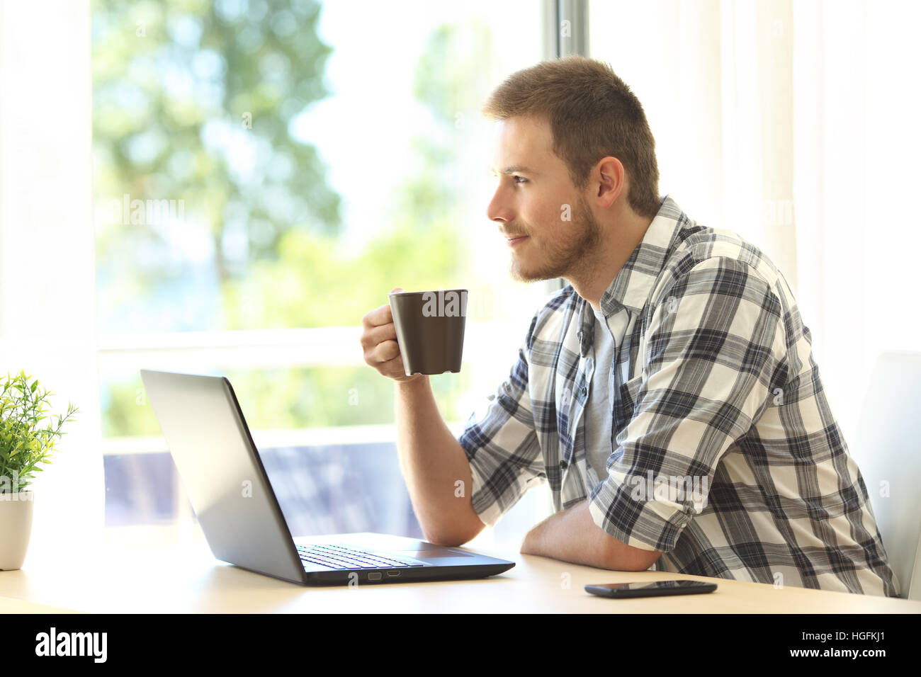 Pensive man with a laptop looking through a window holding a coffee cup at home Stock Photo