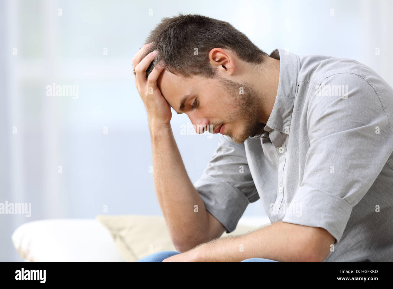 Side view of a sad man with a hand on the head sitting on a couch in the living room at home Stock Photo