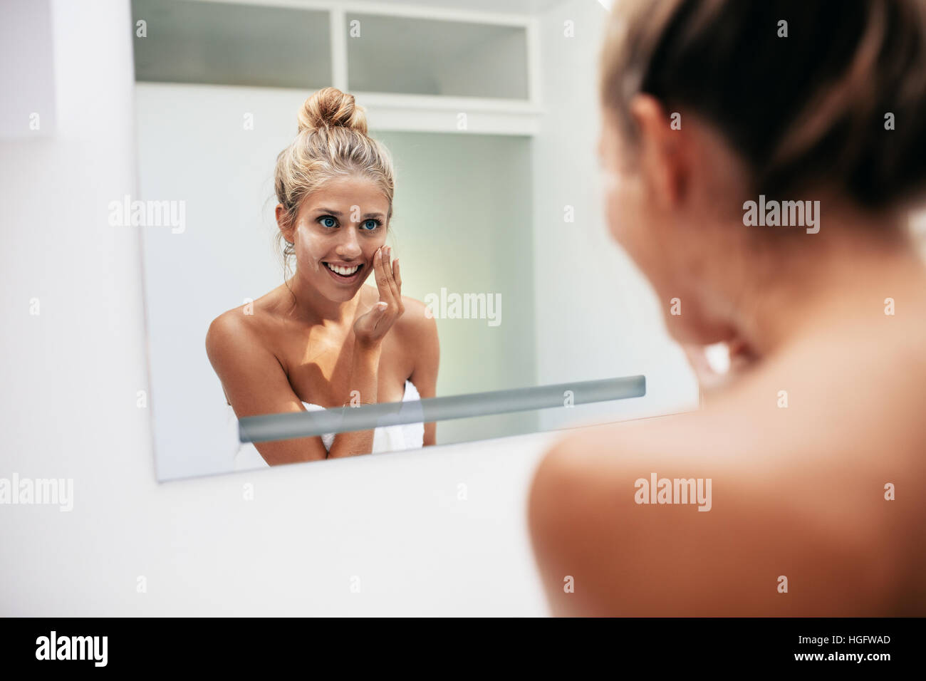 Smiling young woman reflection in mirror applying face cream in bathroom. Caucasian female doing beauty treatment at home. Stock Photo