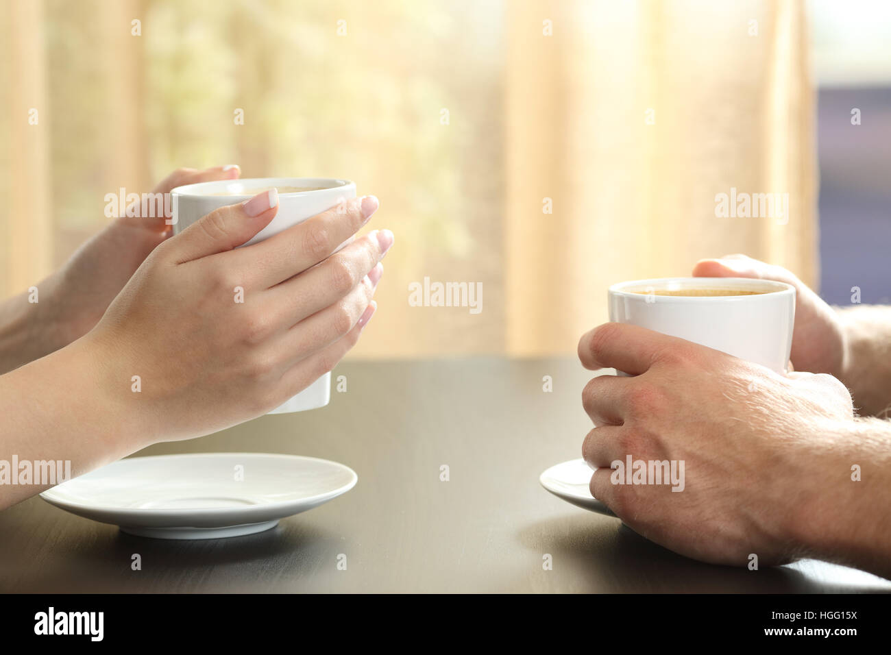 Close up of side view of a couple or friends talking with hands holding coffee cups on a table with a window and curtain in the background Stock Photo