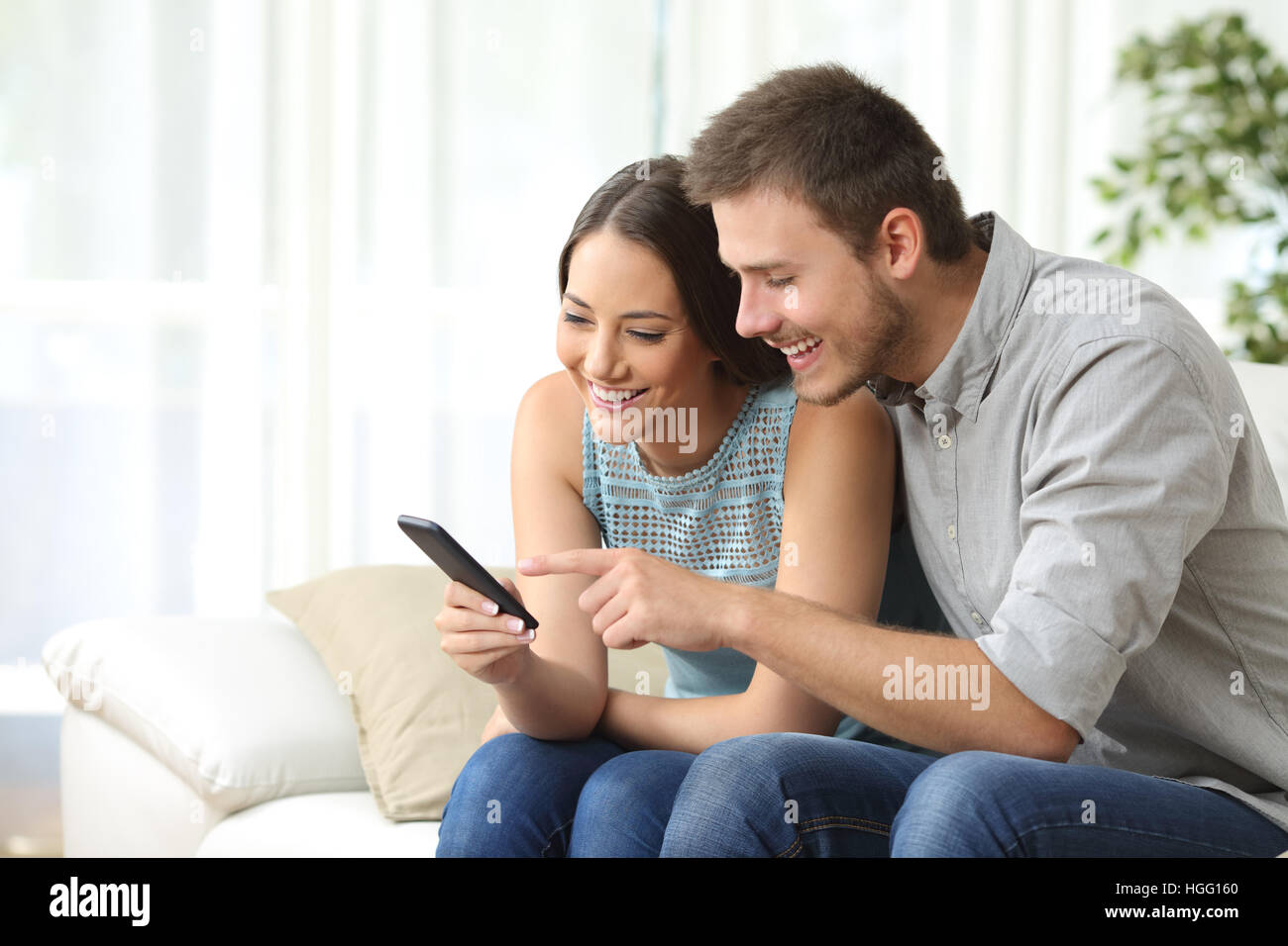 Relaxed couple or friends using a generic mobile phone together sitting on a sofa in the living room at home Stock Photo