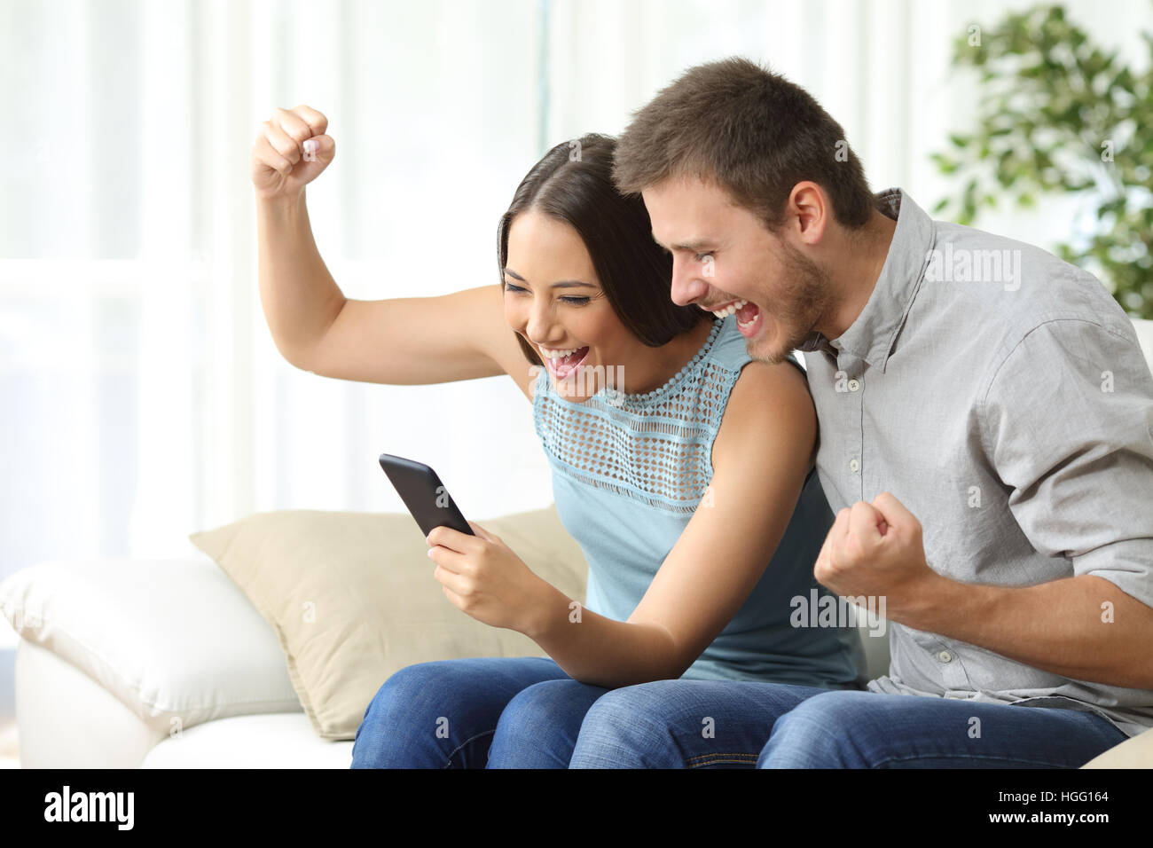 Excited couple watching media content together using a mobile phone sitting on a couch in the living room of a house Stock Photo