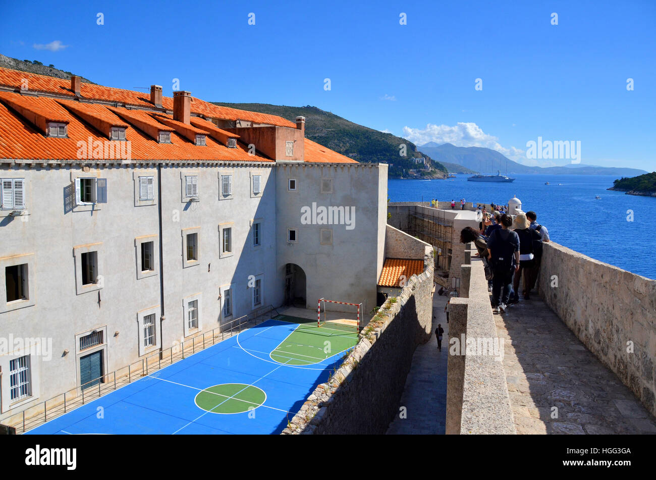 Tourists walk along the wall of the walled city of Dubrovnik, Croatia, passing a netball court belonging to a local school. Stock Photo
