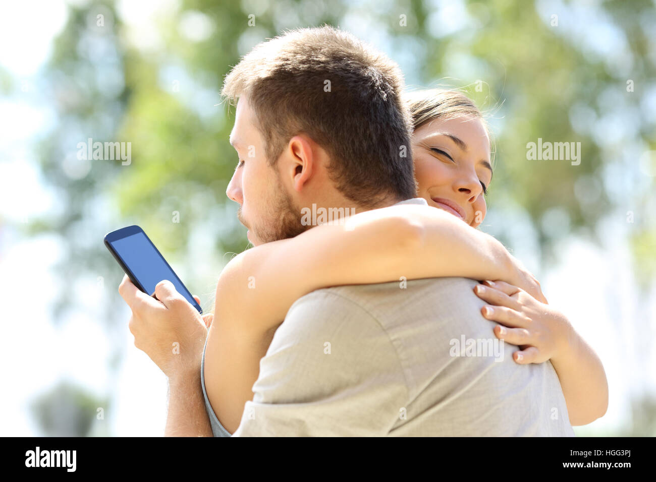 Cheater texting with his other lover on phone and hugging his innocent girlfriend Stock Photo