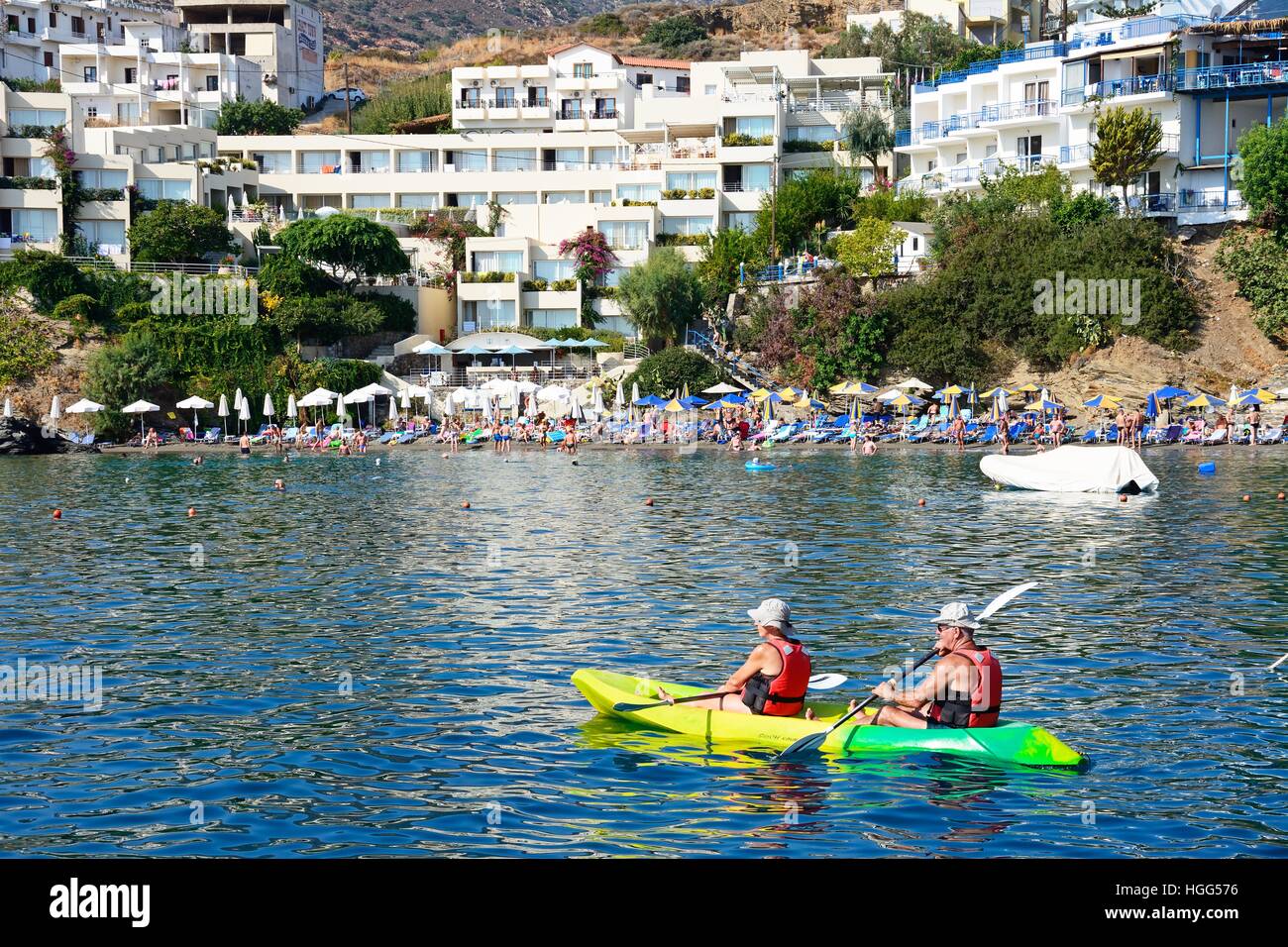 A couple canoeing in the harbour with tourists relaxing on the beach to the rear, Bali, Crete, Greece, Europe. Stock Photo