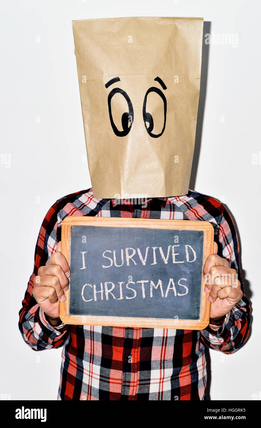 a young caucasian man dressing casual wear with a paper bag in his head shows a chalkboard with the text I survived christmas written in it Stock Photo
