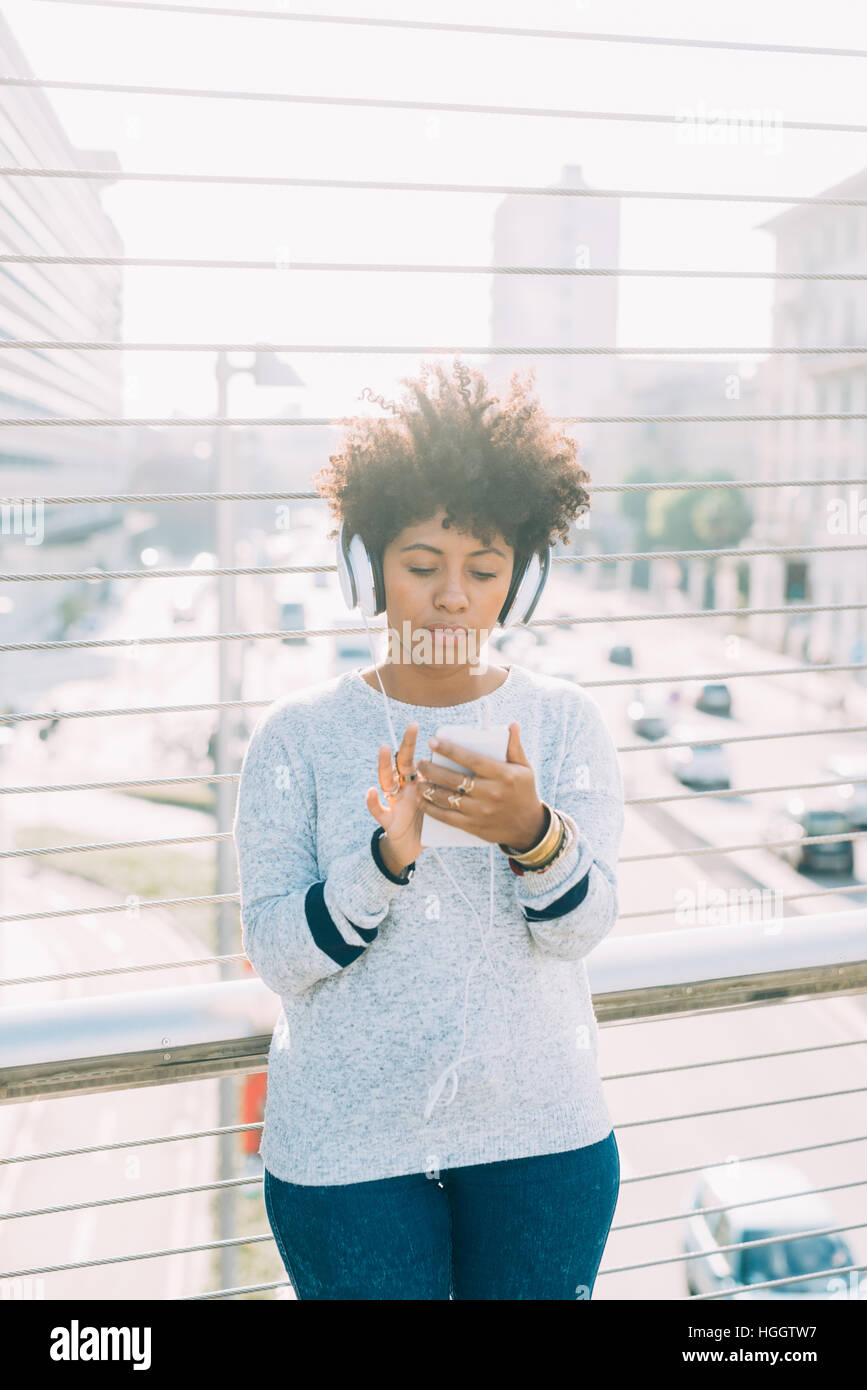 Young beautiful afro woman outdoor in city back light listneing music with head phone and smart phone hand hold, thoughtful - melancholic, music, tech Stock Photo