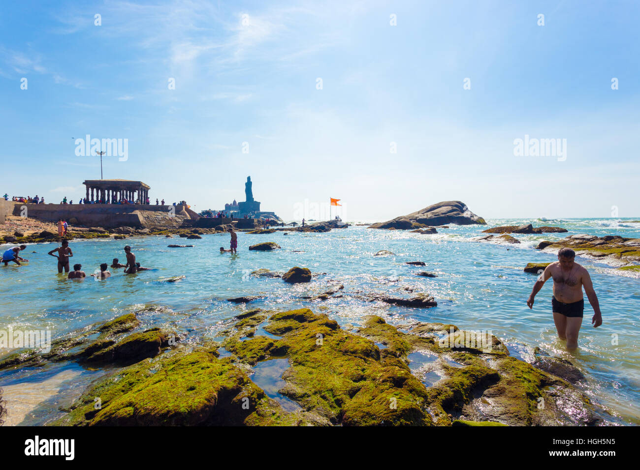 Indian tourists swimming in shallow rocky beach with Thiruvalluvar statue and 16 legged mandap pavilion in background Stock Photo
