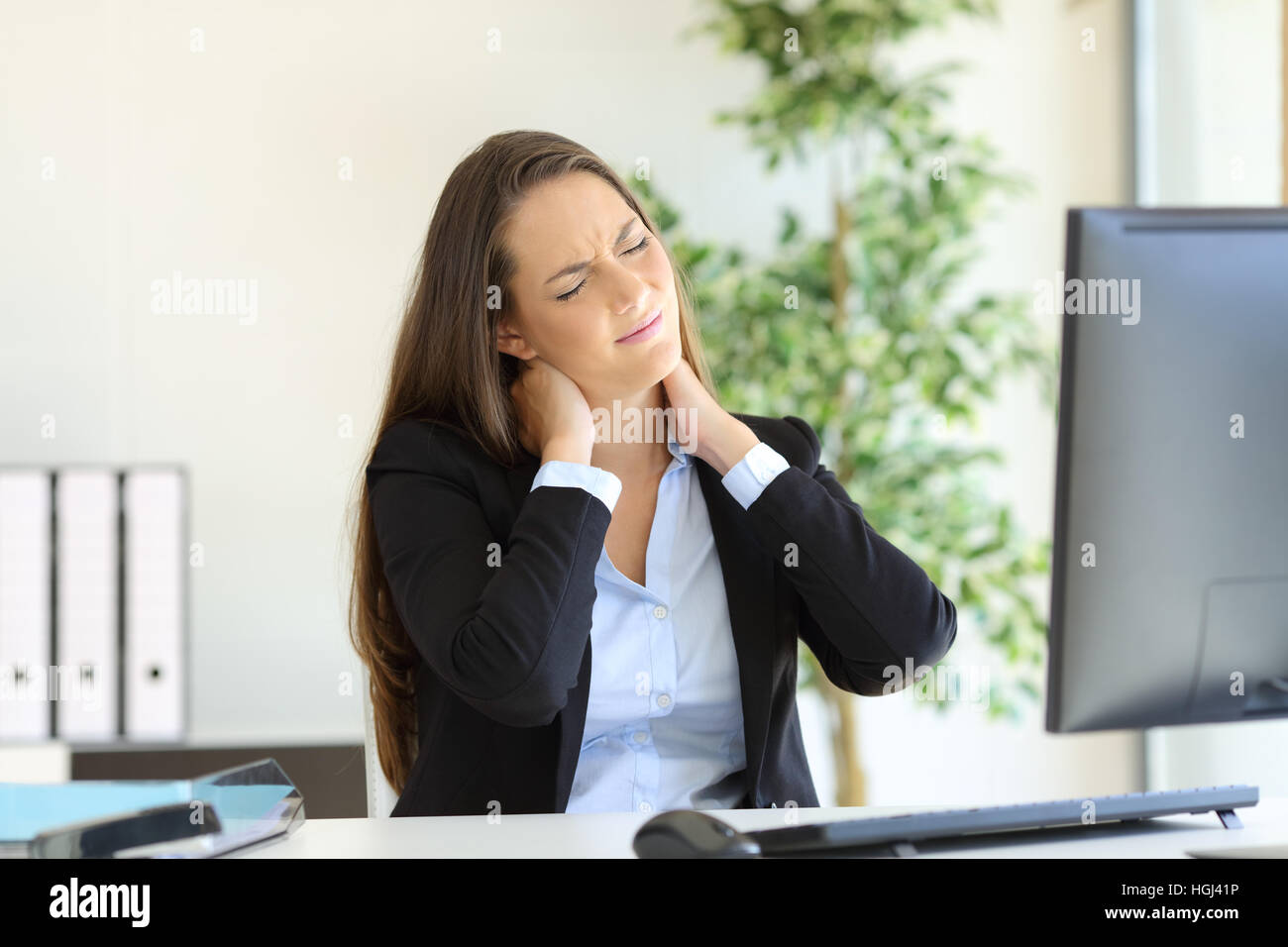 Businesswoman suffering neck pain sitting in a chair while working with a desktop computer in her workplace at office Stock Photo