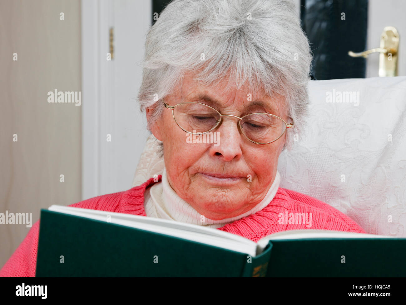 Old aged retired elderly senior woman retiree wearing spectacles enjoying retirement sat relaxing reading a book indoors at home. England UK Britain Stock Photo