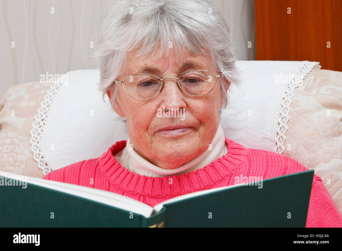 Retired elderly senior woman wearing spectacles sat relaxing in an easy chair and reading a book at home. England, UK, Britain. Stock Photo