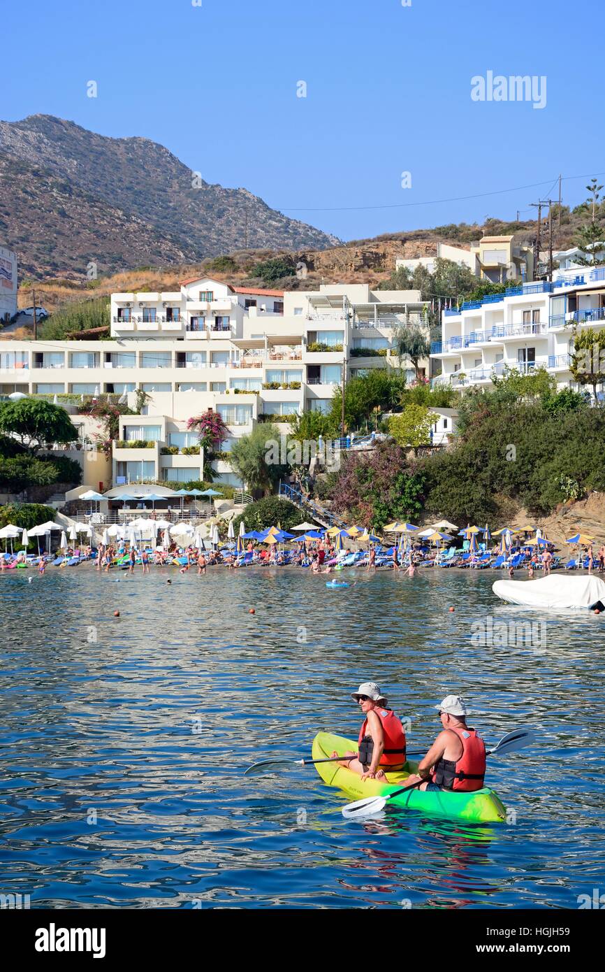 A couple canoeing in the harbour with tourists relaxing on the beach to the rear, Bali, Crete, Greece, Europe. Stock Photo