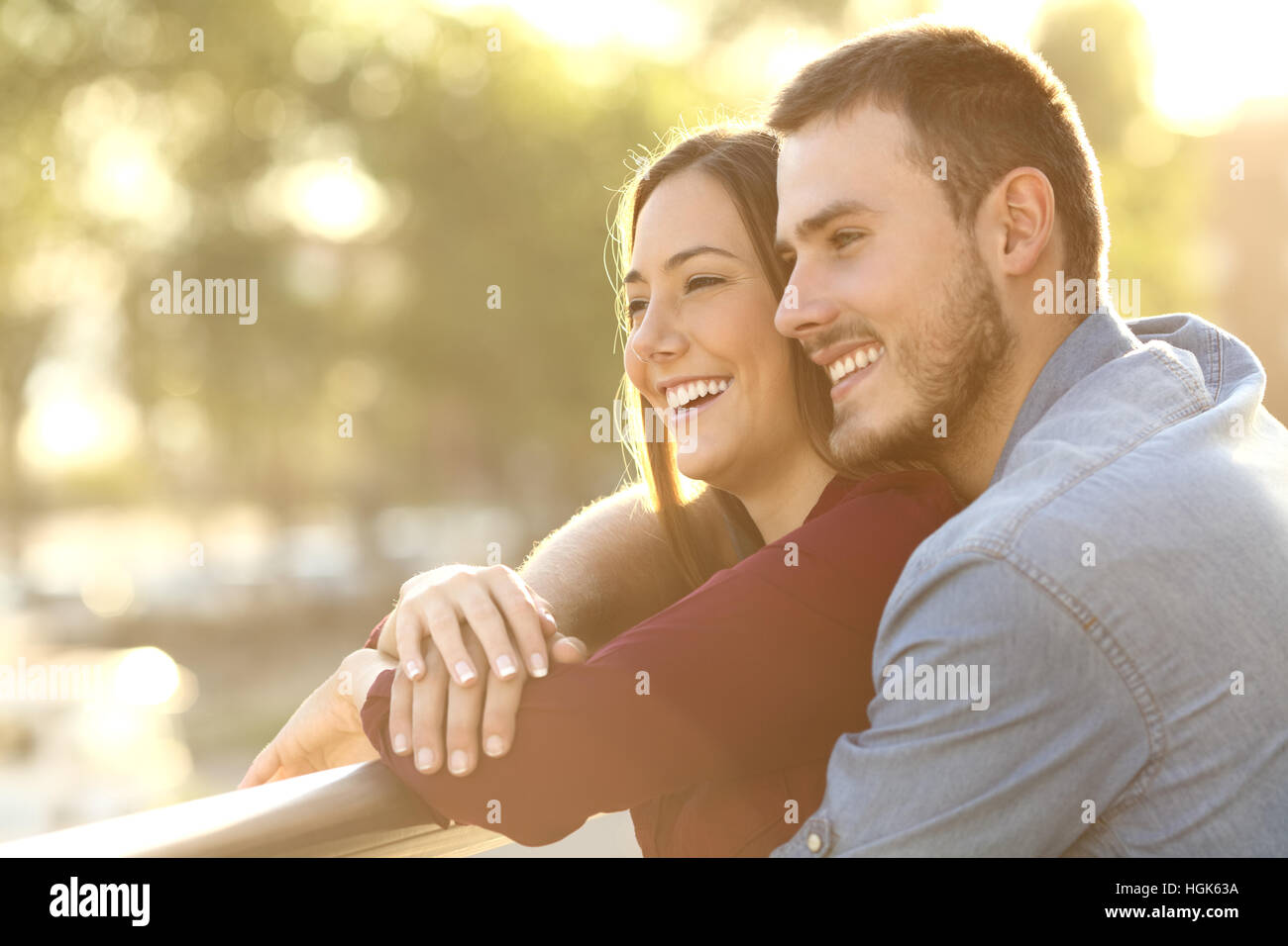Enamored couple embracing and looking away enjoying views at sunset in a balcony with a warm light in the background Stock Photo