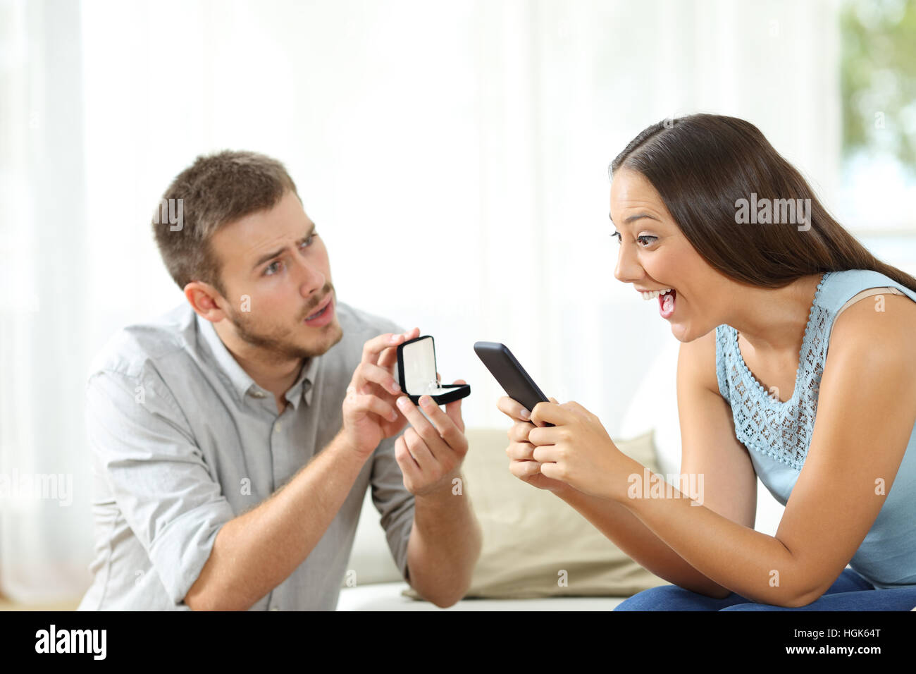 Amazed woman using a smart phone ignoring a marriage proposal of her worried boyfriend at home Stock Photo