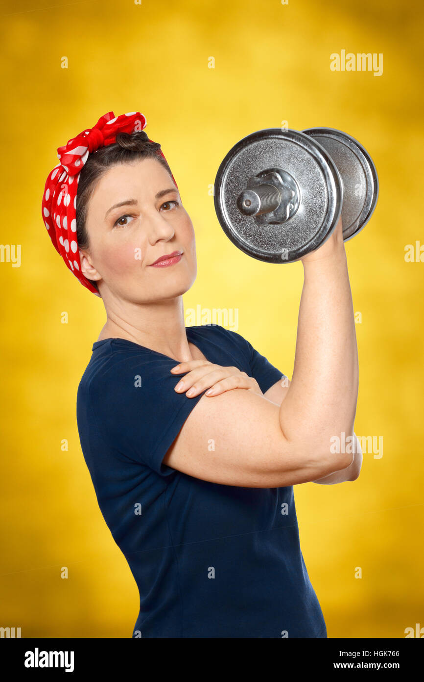 Smiling woman with red kerchief and blue t-shirt lifting a heavy dumbbell, yellow background, american icon Rosie the Riveter Stock Photo