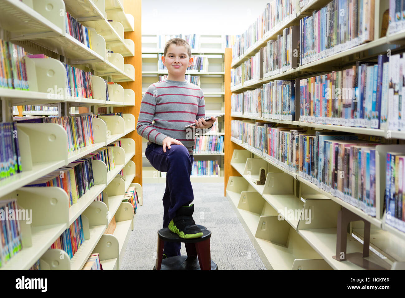 Happy boy in library surrounded by book shelves with foot on stool Stock Photo