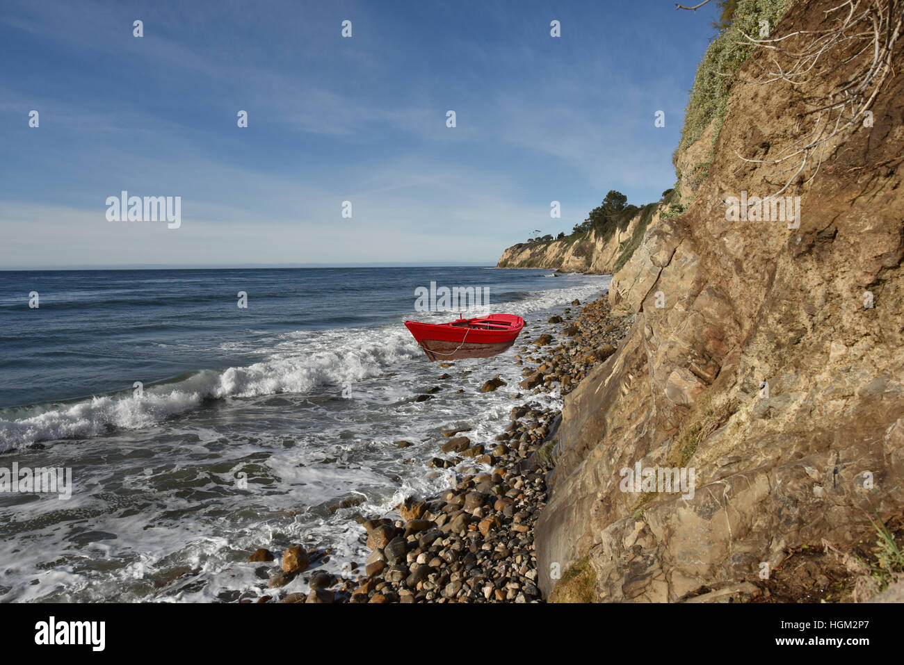 Red rowboat in the surf on a rocky beach beneath a cliff. Stock Photo