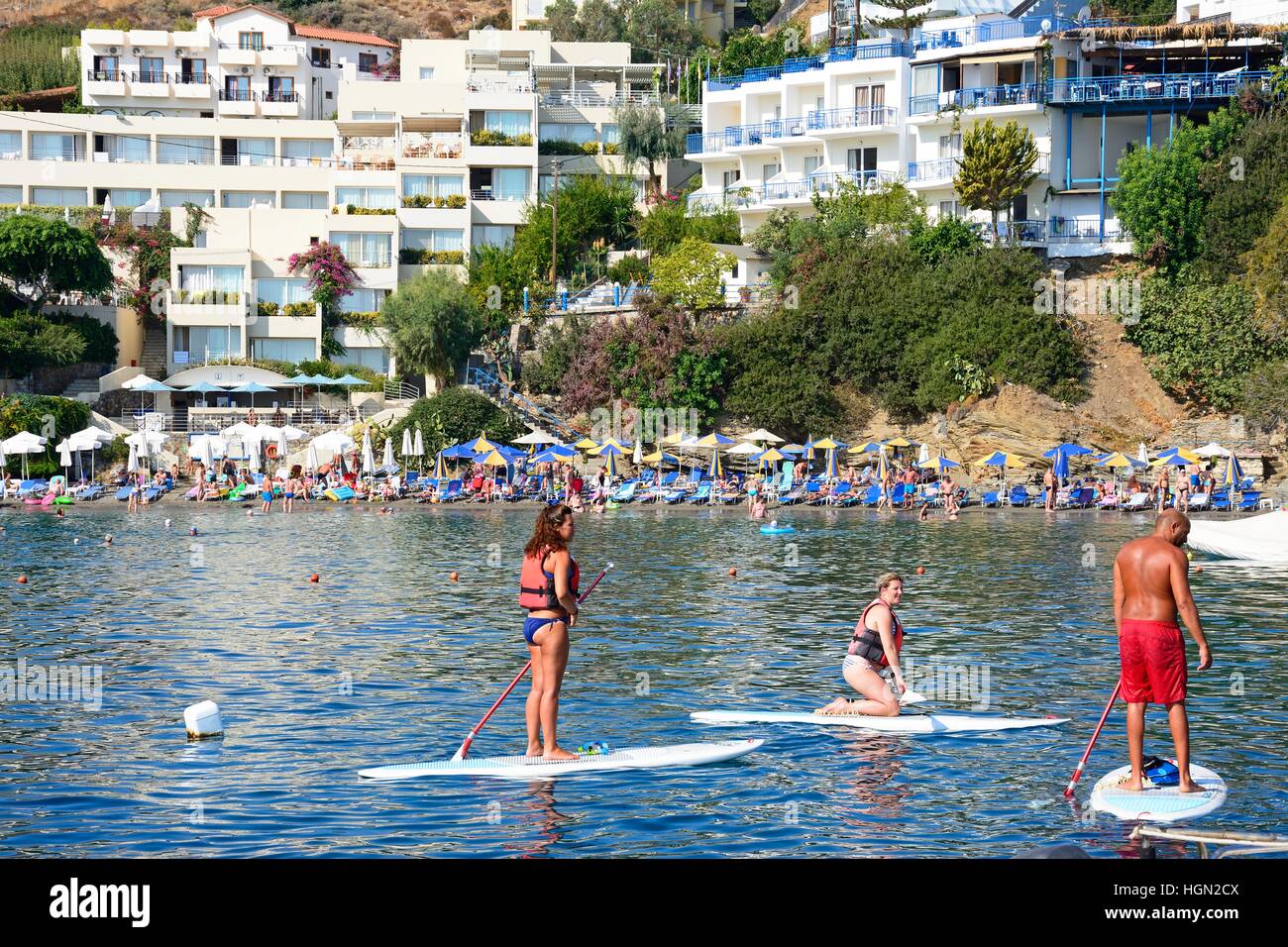 Holidaymakers on paddle boards with tourists relaxing on the beach to the rear, Bali, Crete, Greece, Europe. Stock Photo