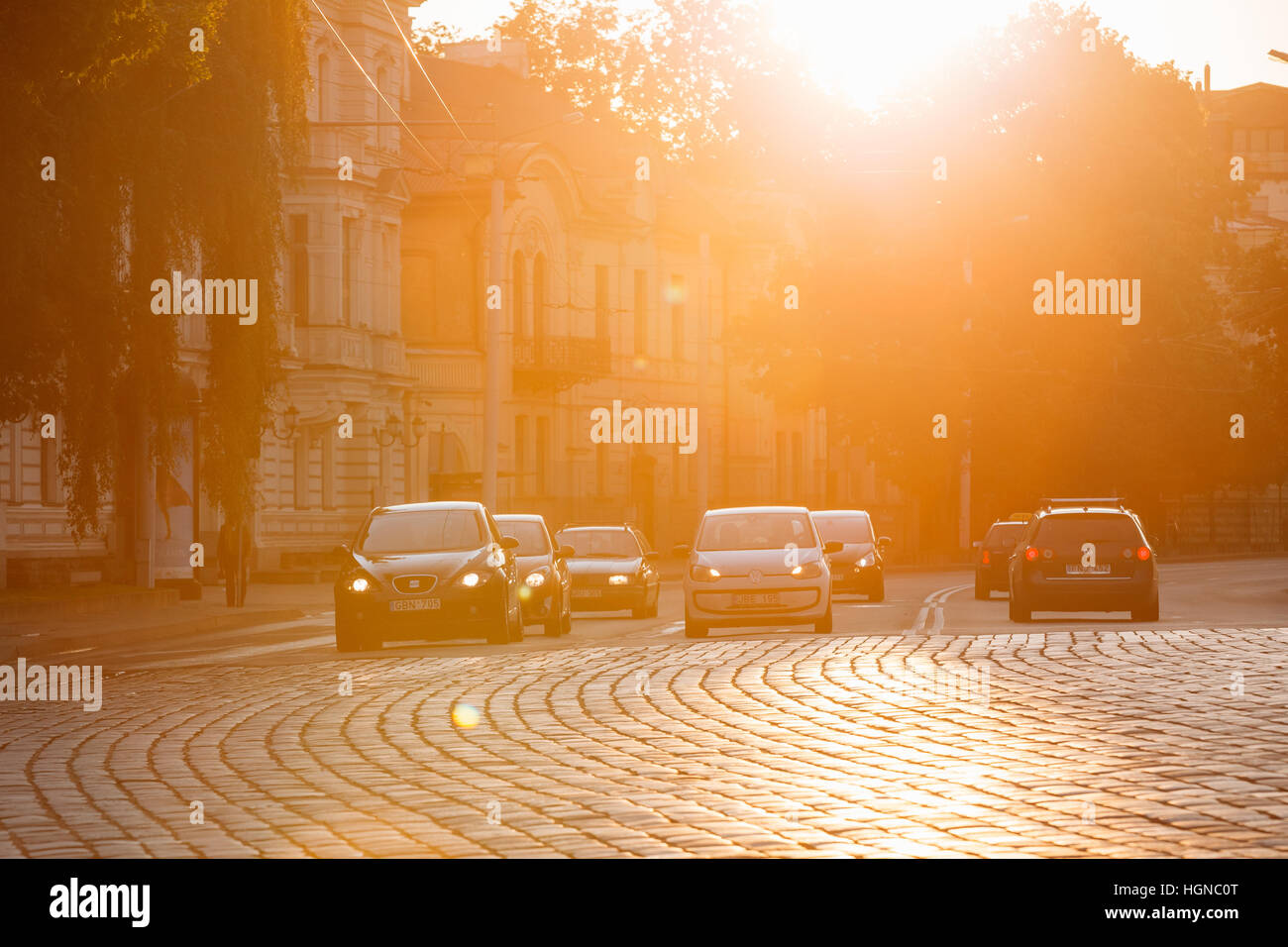 Vilnius, Lithuania  - July 8, 2016: Traffic On Zygimantu Street In Old Town. Moving Cars With Luminous Headlights On The Paved Road In Yellow Sunlight Stock Photo