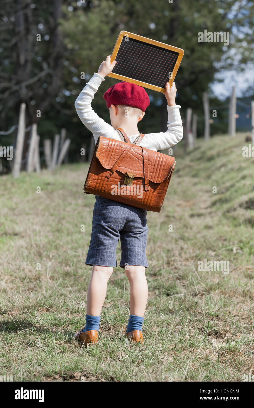 Young school boy in vintage clothing and red beret hat with leather school bag on his back hold up a small blackboard over his head (copy space) Stock Photo