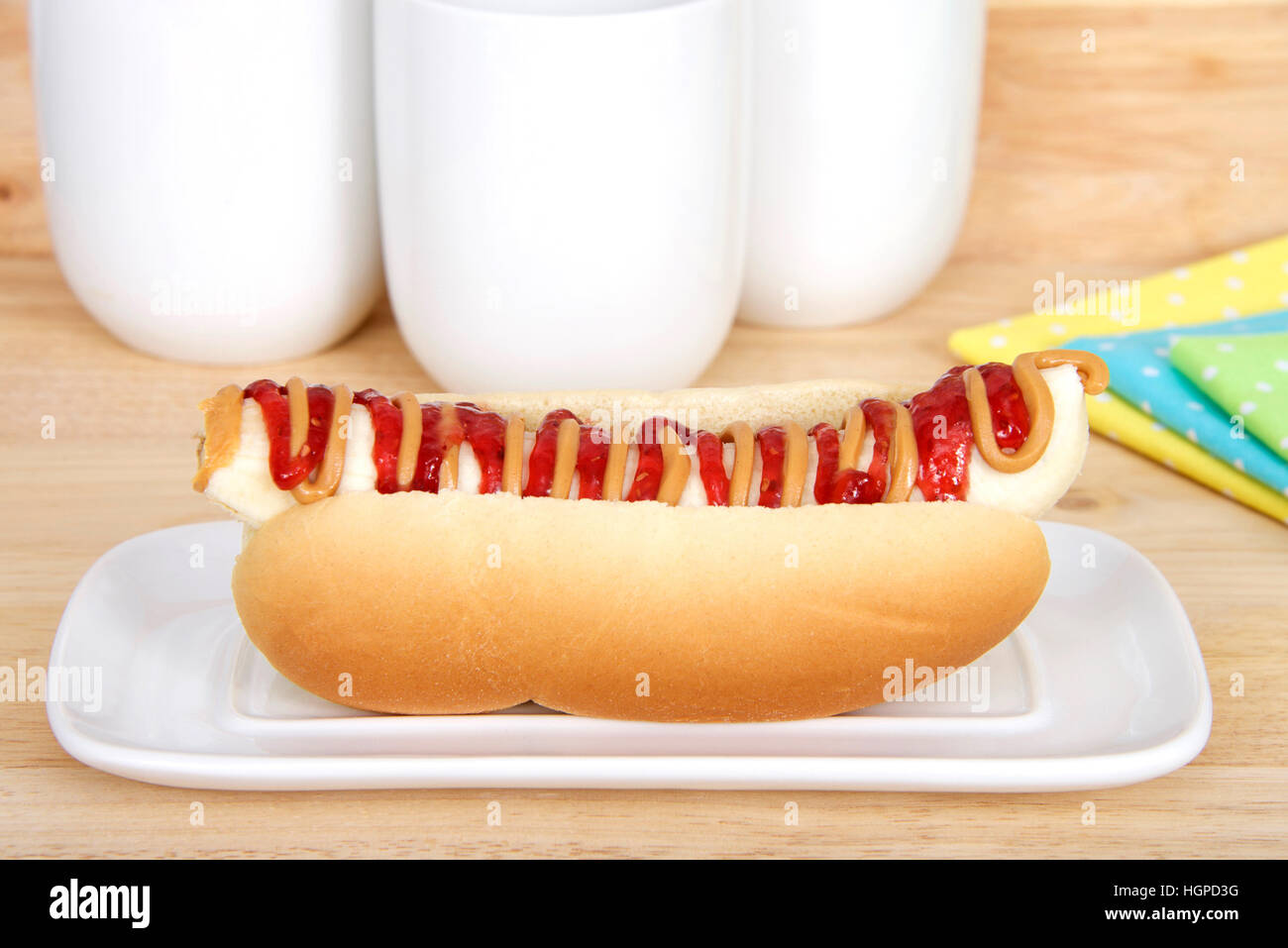 Banana with peanut butter and jelly in a hot dog bun served on a white rectangular plate, yellow, blue, green polka dot napkins and cups in background Stock Photo