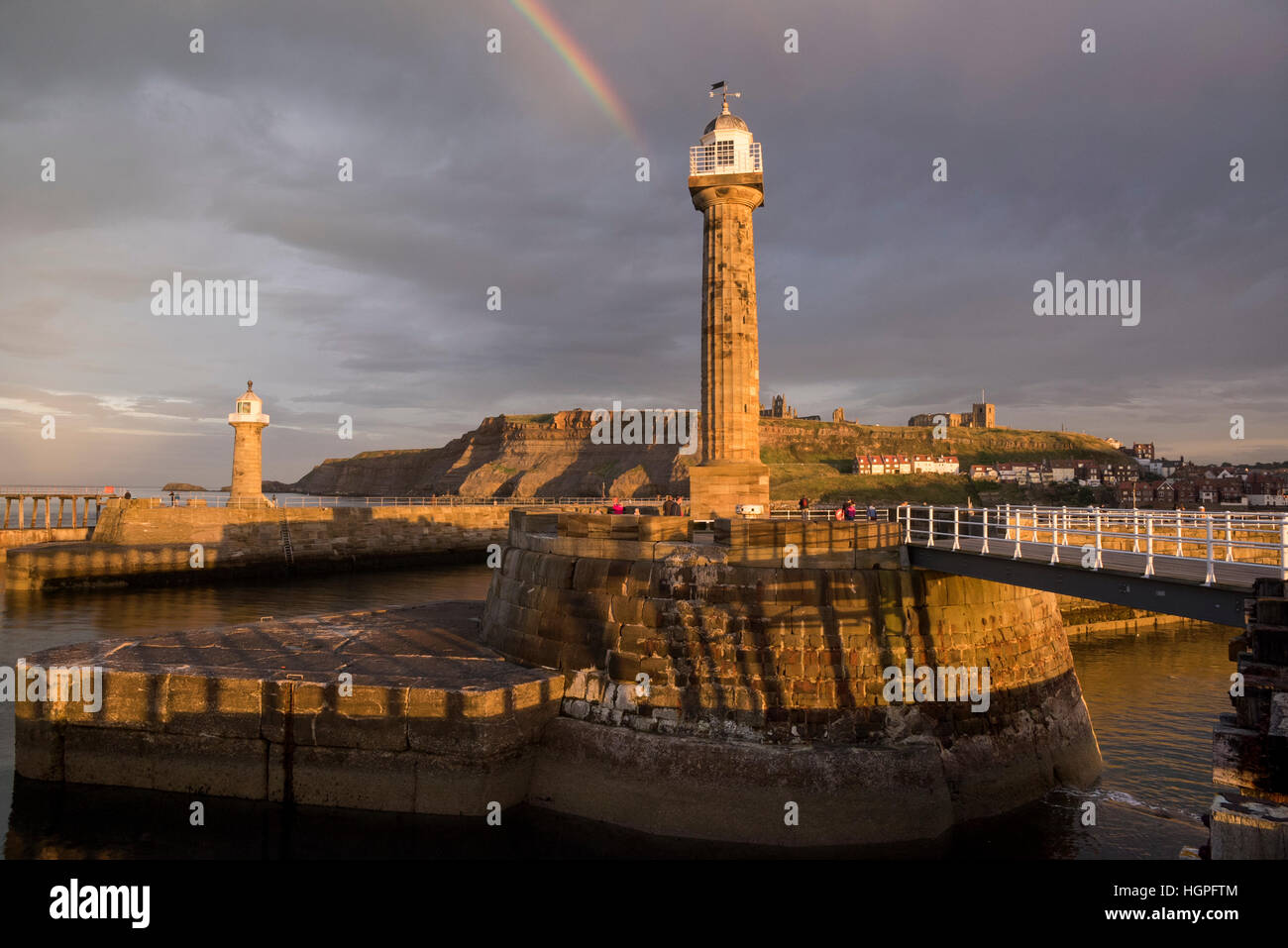 Rainbow and dark sky over sunlit lighthouses, harbour entrance, the Abbey and St. Mary's Church - Whitby, North Yorkshire, GB. Stock Photo