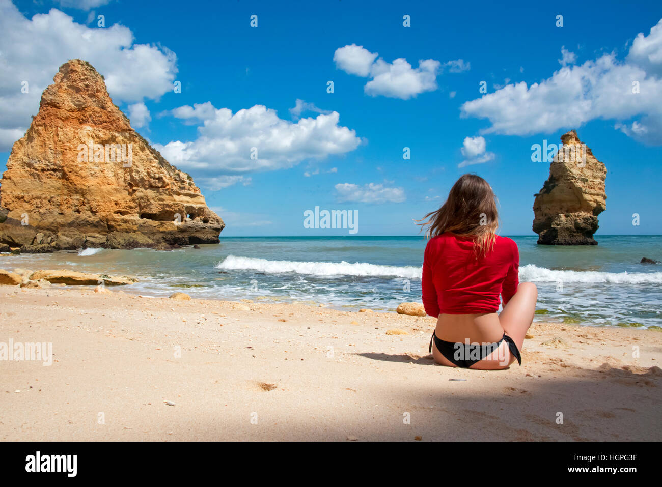 Young girl sitting in the white sand on the beach wearing black Brazilian bikini and red top Stock Photo
