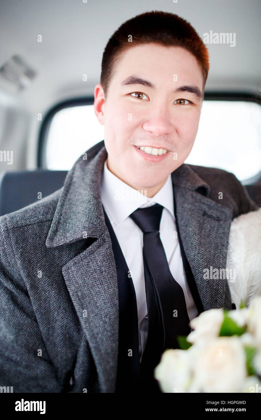 The groom in a winter coat sitting in the car with wedding bouquet in hands. Positive portrait, looking into the camera. Stock Photo