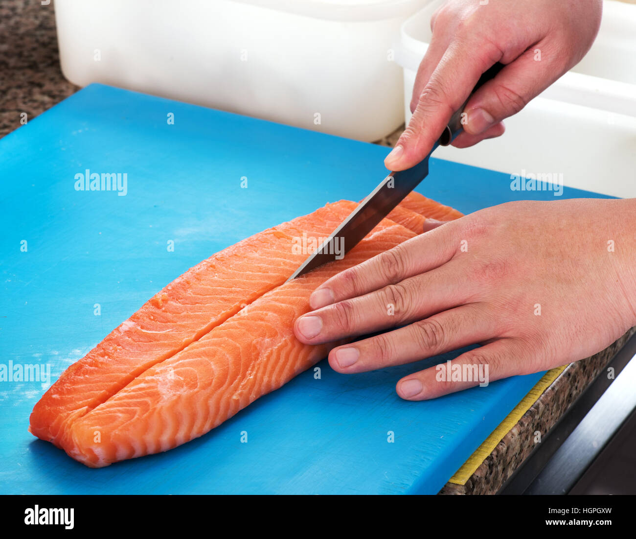 Sushi chef slicing a raw fresh salmon fillet with a sharp knife on a blue cutting board in a restaurant, close up of his hands Stock Photo