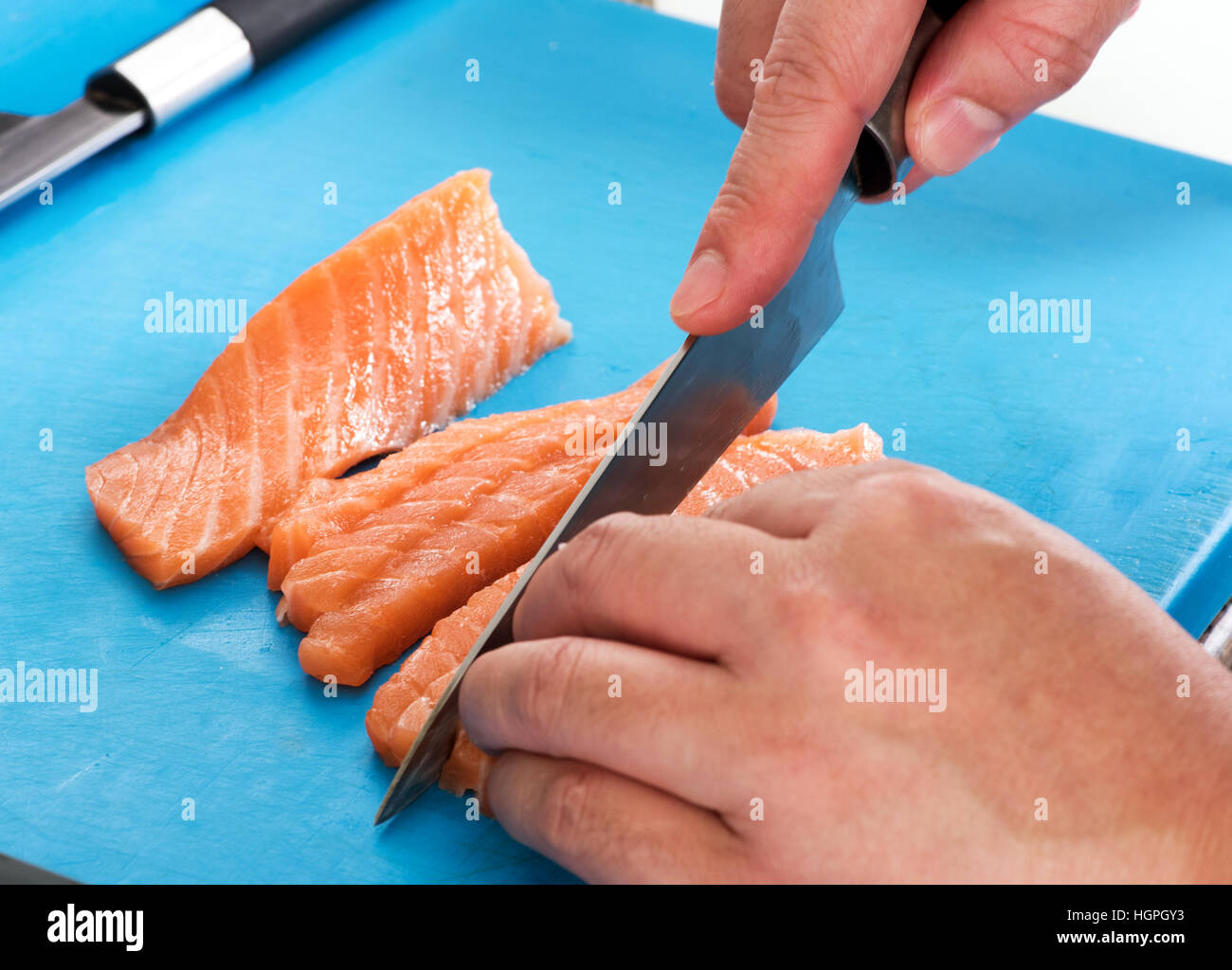 Chef slicing raw fresh salmon fillet with a sharp knife in a Japanese sushi bar , close up view on his hands and the fish on a blue cutting board Stock Photo