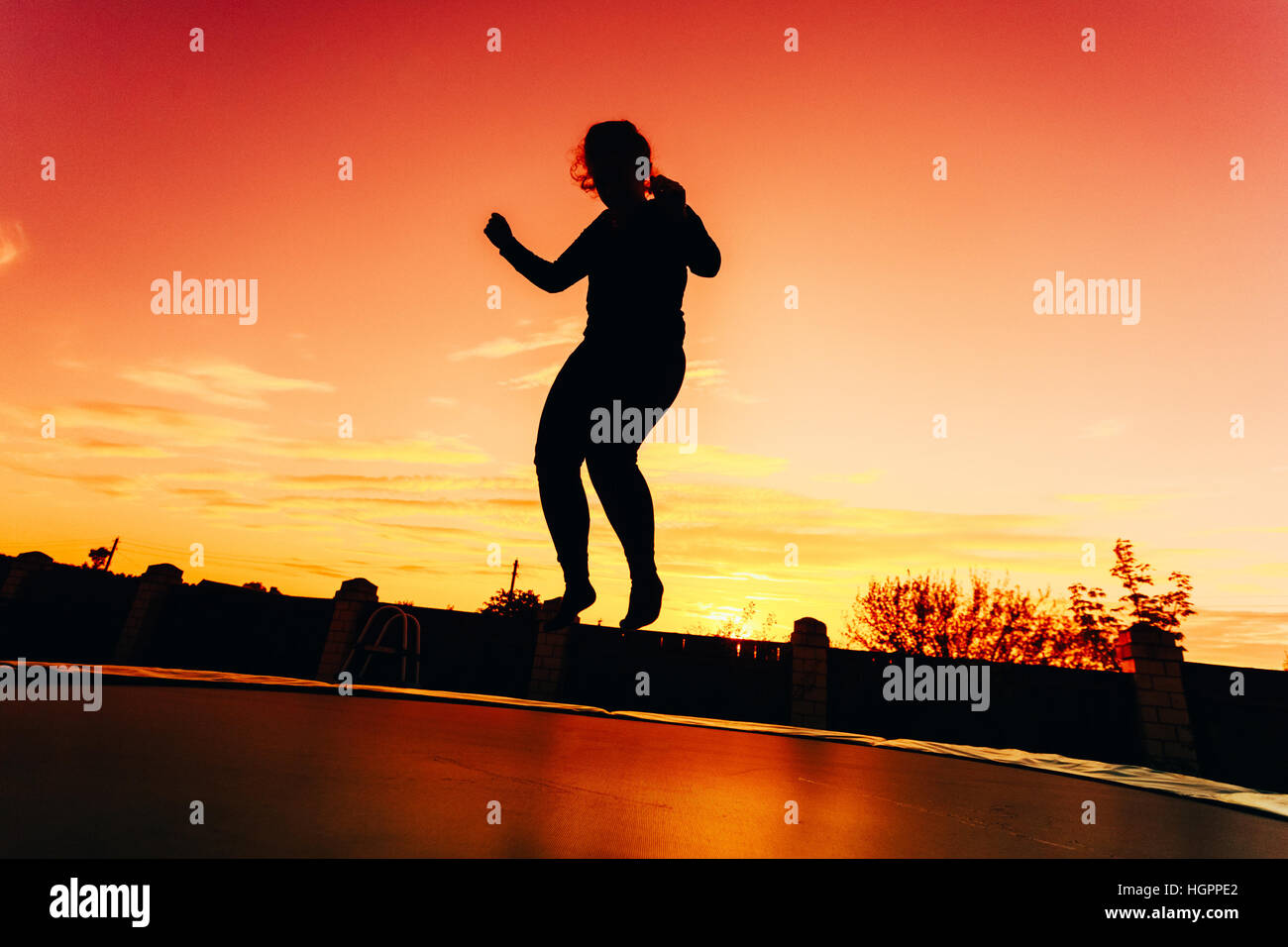 Silhouette Of Beautiful Plus Size Young Woman Girl Jumping On Trampoline On Evening Red, Orange Colorful Sunset Sky Background Stock Photo