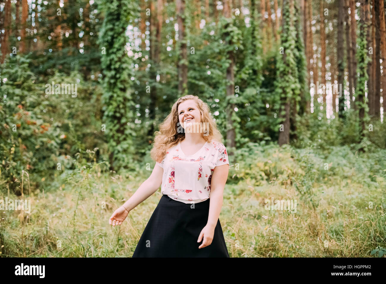 Young Pretty Plus Size Caucasian Happy Smiling Laughing Girl Woman With Wavy Brown Long Hair In White T-Shirt, Waltz Round In Summer Green Forest. Stock Photo