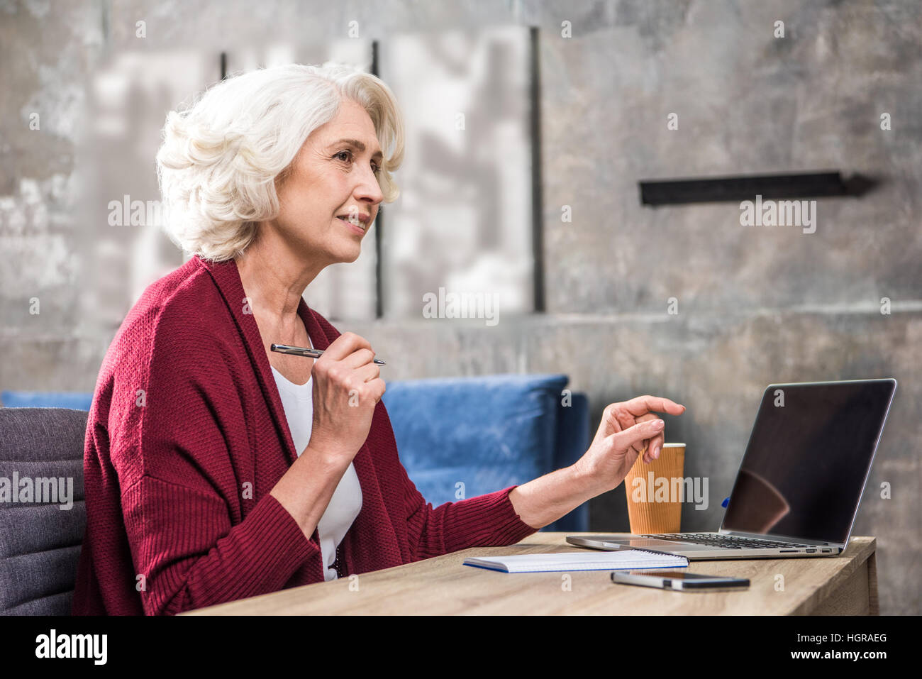 Attractive smiling senior woman freelancer holding pen sitting at the table with laptop Stock Photo
