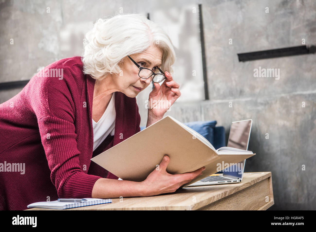 Attractive senior woman in eyeglasses reading book while sitting at desk with laptop Stock Photo