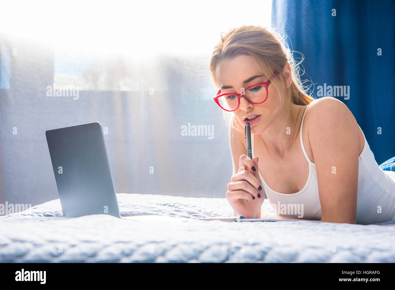 Beautiful teenage girl in eyeglasses making notes in notebook while lying on bed Stock Photo