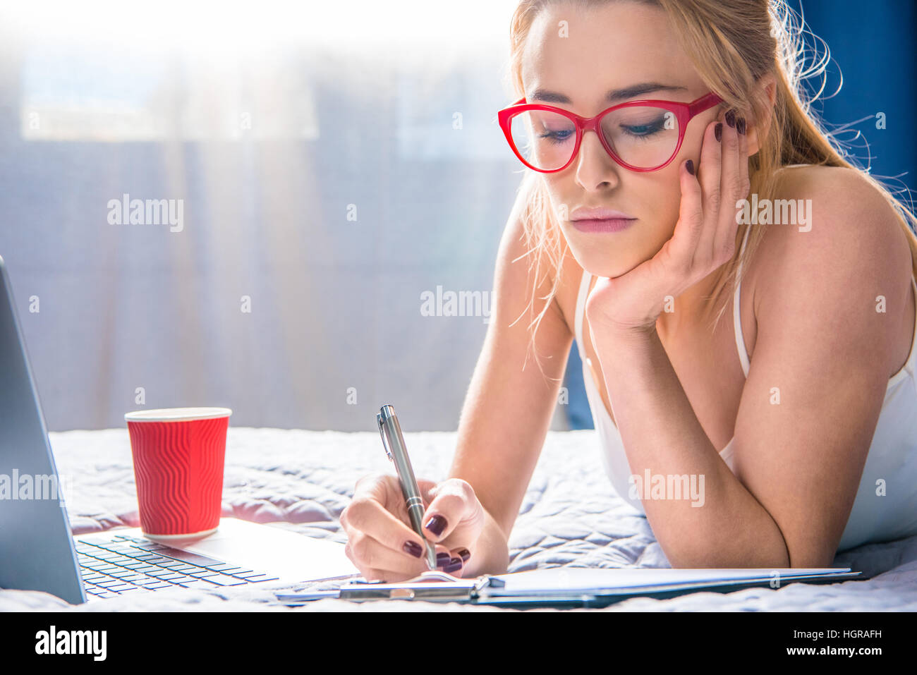 Beautiful teenage girl in eyeglasses making notes in notebook while lying on bed Stock Photo