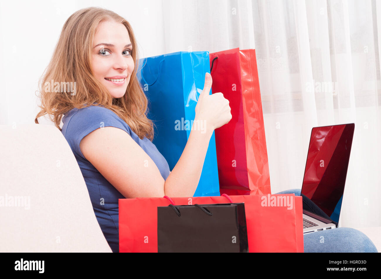Woman sitting on couch making online order showing like gesture with shopping bags around Stock Photo