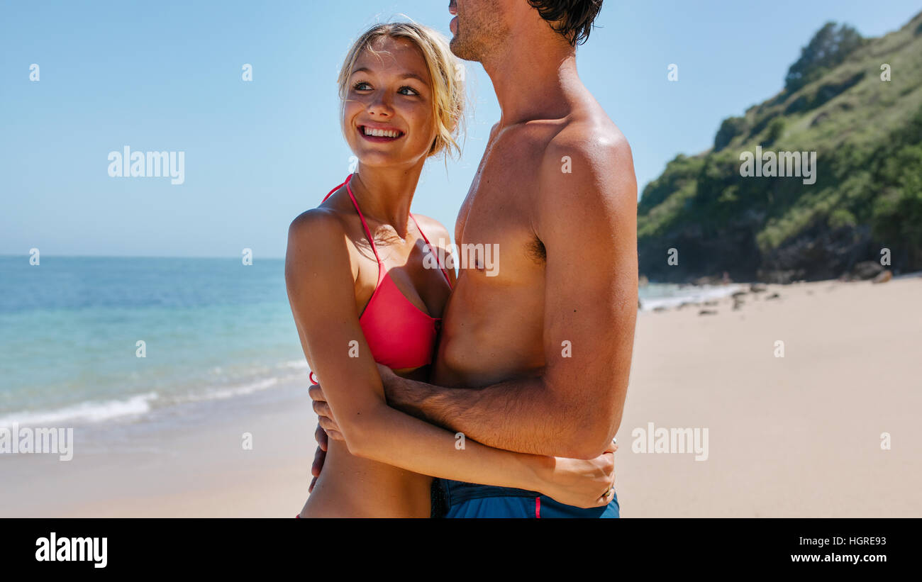 Portrait of happy young couple standing together on the beach. Beautiful young woman embracing her boyfriends and looking away smiling. Stock Photo