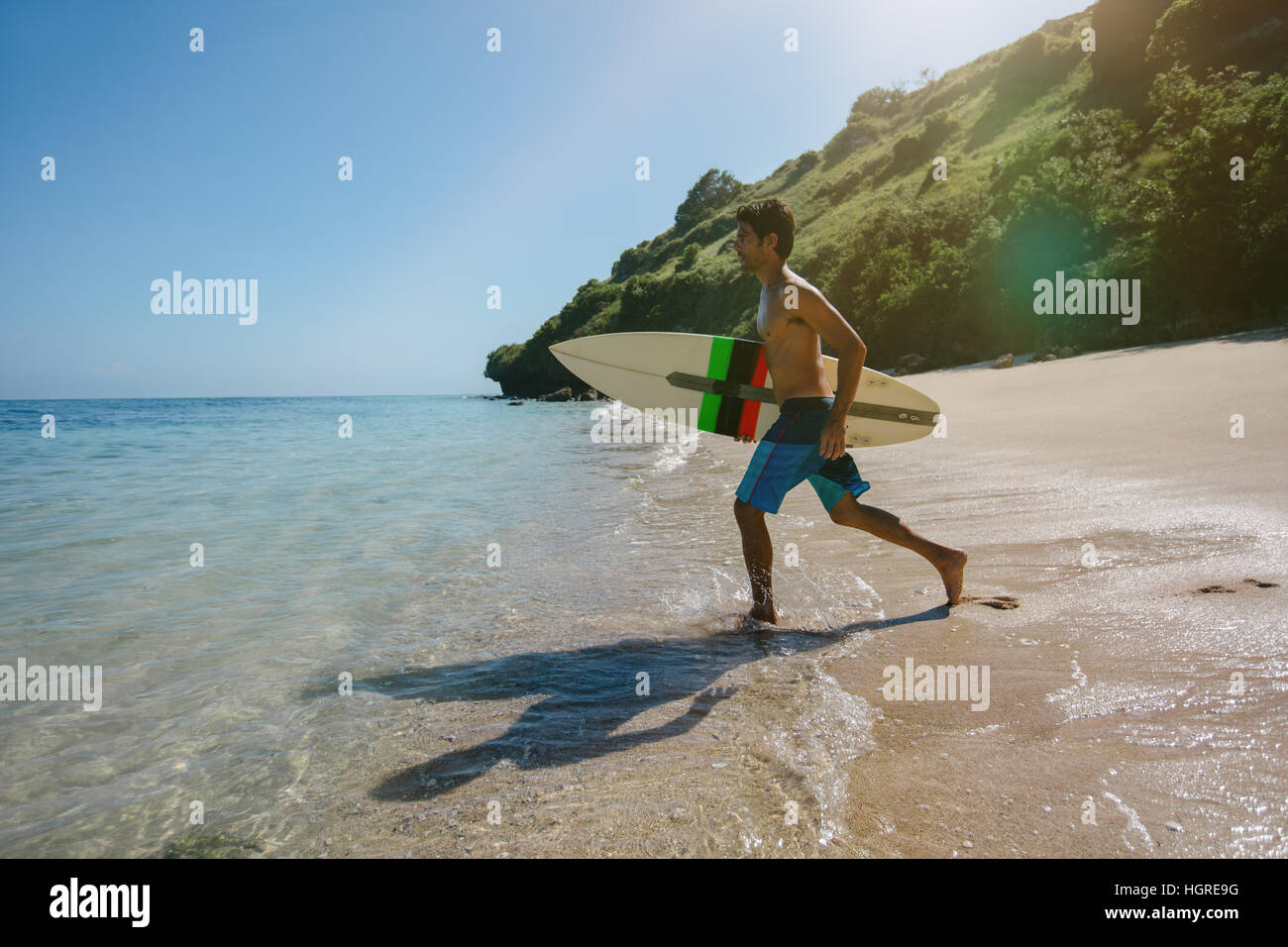 Shot of handsome young man with surfboard going for surfing in the sea. Surfer carrying his surfboard running on the beach. Stock Photo