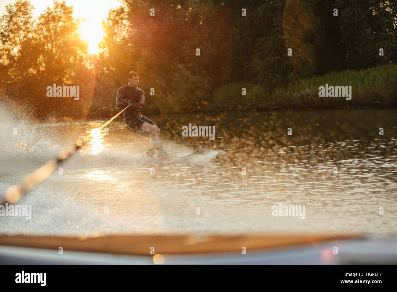 Man riding wakeboard on wave of motorboat in lake at sunset. Male athlete in action, water skiing on lake. Stock Photo