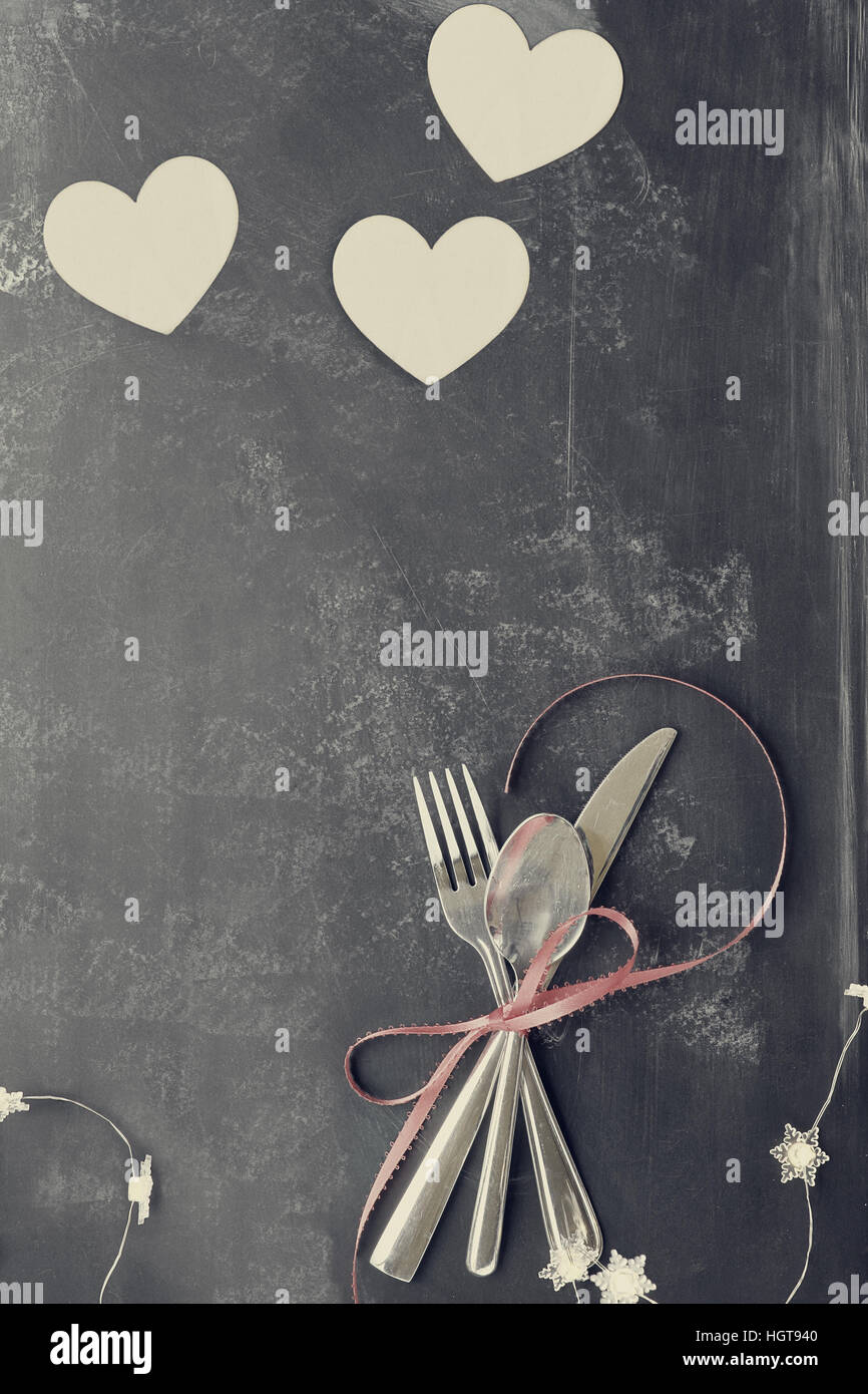 Top view of Valentines day dinner table setting with cutlery over a rustic blackboard background with wooden cut out hearts and fairy lights. Stock Photo