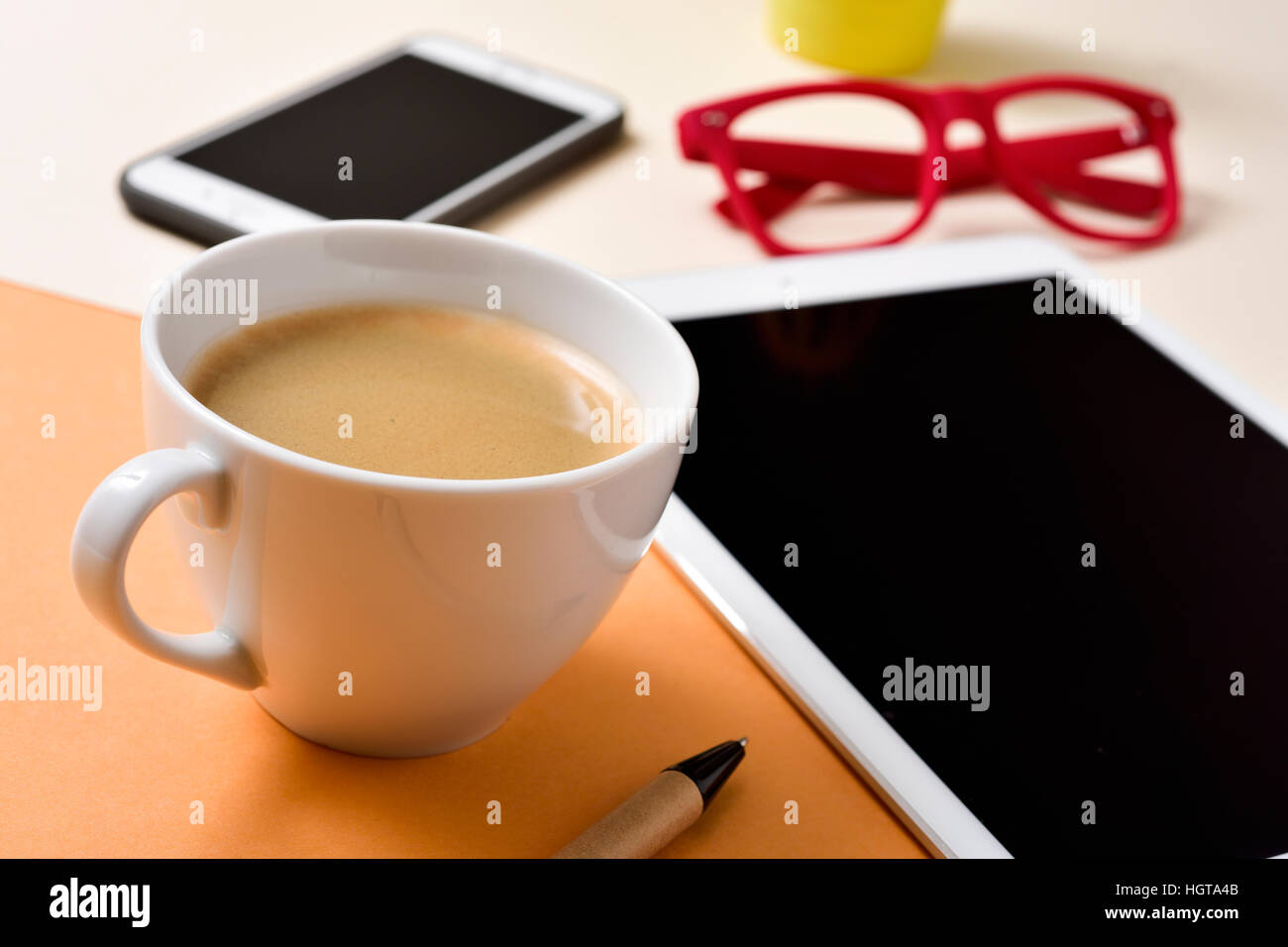 closeup of an office desk full of things, such as a cup with white coffee, a pen, a tablet, a smartphone and a pair of red eyeglasses Stock Photo