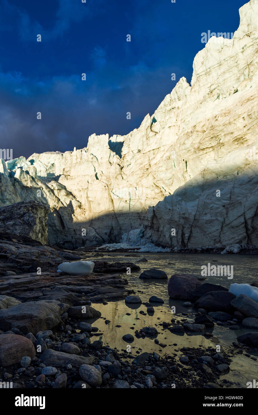 The setting sun illuminates the sheer ice cliff of a glacier fracture zone with a river at it's base. Stock Photo