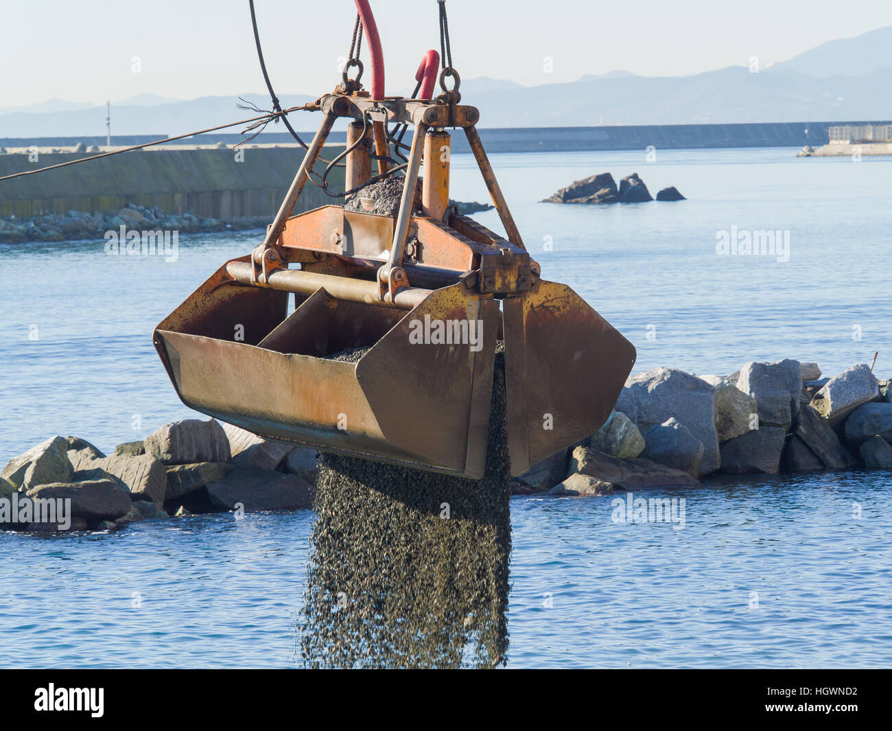 Dredge Clamshell Bucket unloading gravel in the water of a port next to the shore to replenish a beach Stock Photo