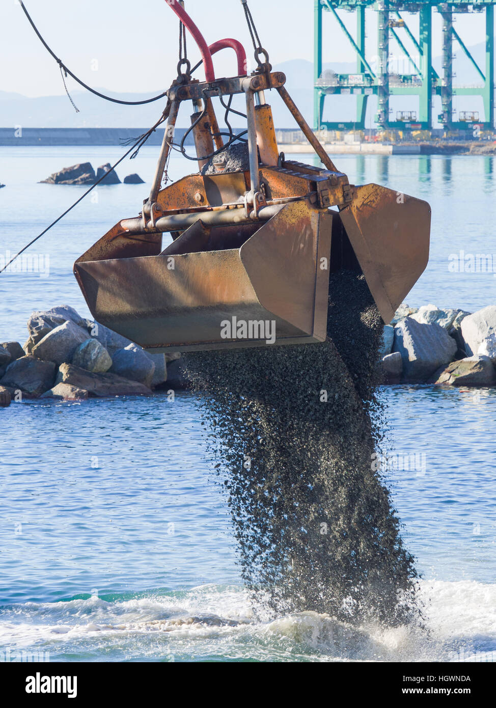 Dredge Clamshell Bucket unloading gravel in the water of a port next to the shore to replenish a beach Stock Photo
