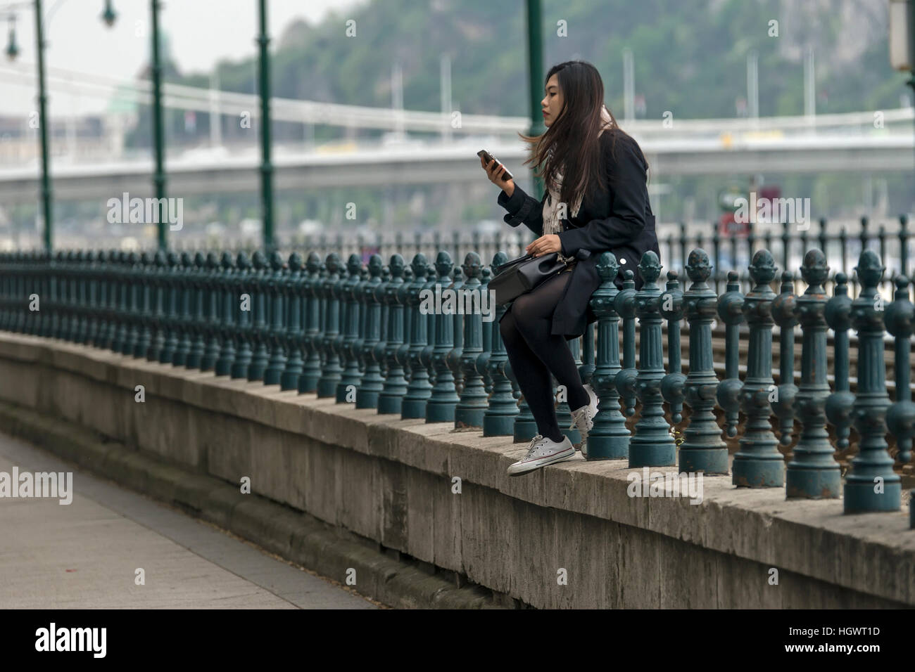 Budapest, Hungary - April 11,2016: A young Chinese girl sitting on the fence and play with mobile phone. Stock Photo
