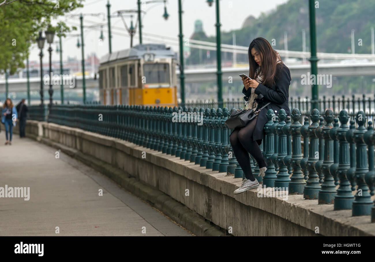 Budapest, Hungary - April 11,2016: A young Chinese girl sitting on the fence and play with mobile phone. Stock Photo
