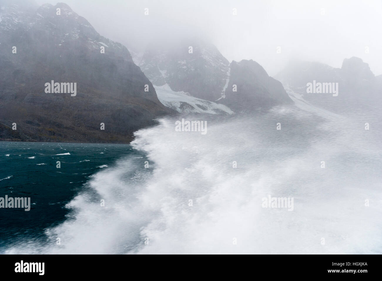 A boats passage through rough seas sends waves and spray into the air beneath jagged mountains. Stock Photo
