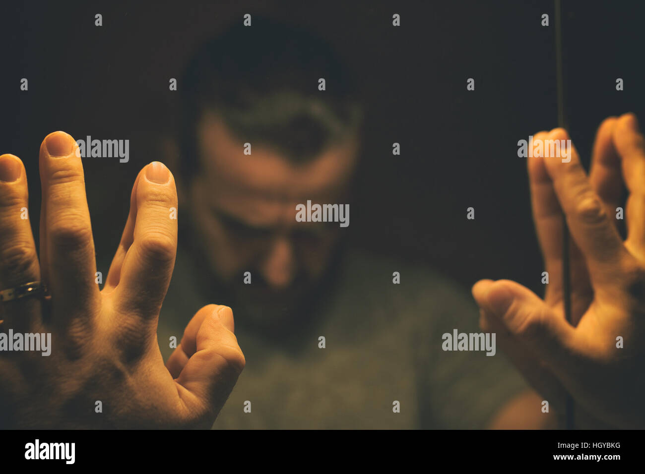 Man in despair with raised hands and bowed hand, in a low light room looking in front of mirror Stock Photo