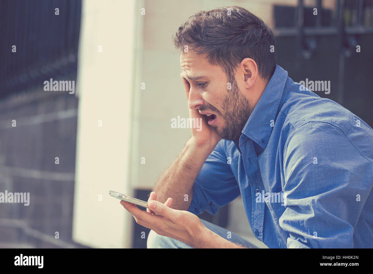 Desperate sad young man looking at bad text message on mobile phone sitting outside office on summer day. Human emotions Stock Photo